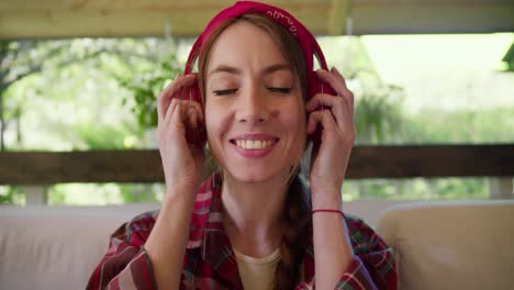 shooting close up a girl in a red plaid shirt puts on red wireless headphones listening to music and shaking her head sitting on a sofa in a gazebo in nature