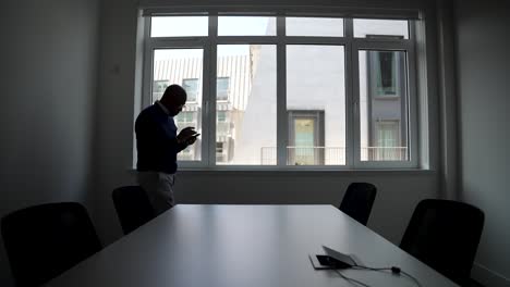 professional male pacing beside a conference table in a modern office setting, attentively checking his tablet device with large windows behind him showcasing an urban cityscape