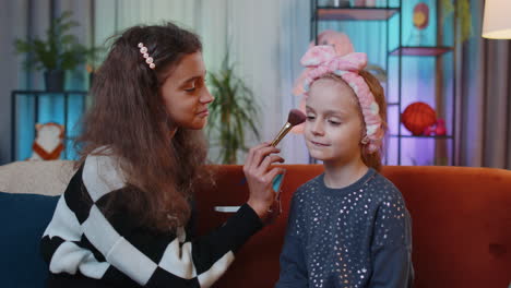 teenage child helps to do face cosmetic makeup her little sibling sister kid girls at home play room