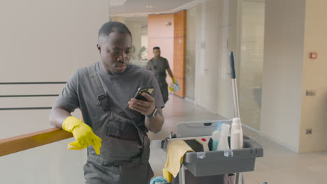 cleaning man wearing gloves leaning on the stair railing while looking at his smartphone next to the cleaning cart 1