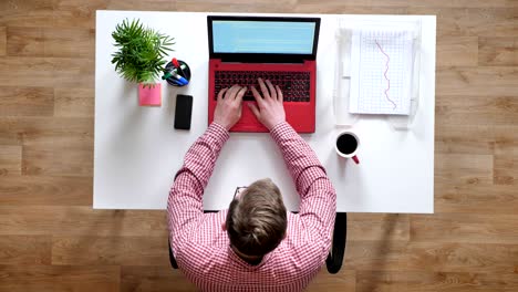 young man in glasses working and typing on laptop, somebody walking around, topshot, sitting behind desk with coffee