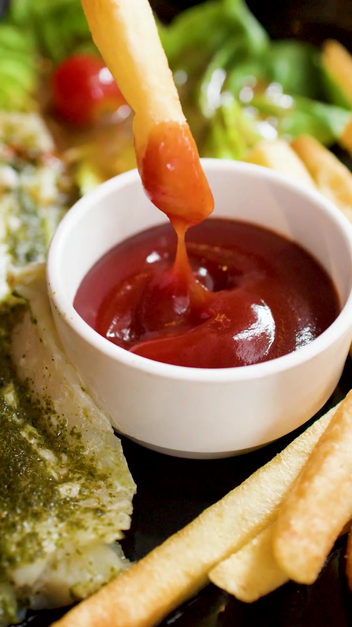 Nahaufnahme einer Hand, die eine goldene Pommes in eine Schale Ketchup taucht.