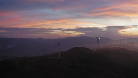 Increíble-Imagen-De-Un-Dron-Volando-A-Través-De-Montañas-Brumosas-Cerca-De-Una-Flota-De-Turbinas-Eólicas-Durante-Una-Colorida-Puesta-De-Sol