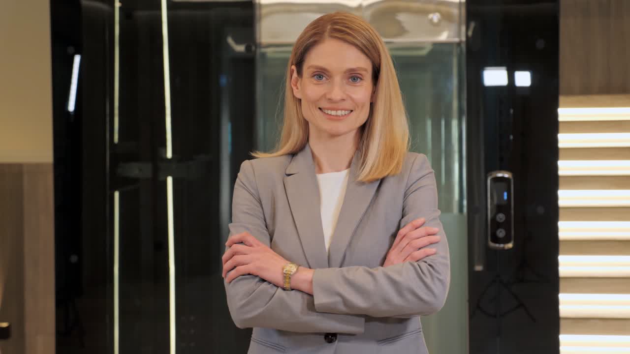 Smiling confident young adult european business woman looking at camera standing in the hall near elevator. Happy blonde professional lady with white teeth pretty face crossing arms close up portrait.