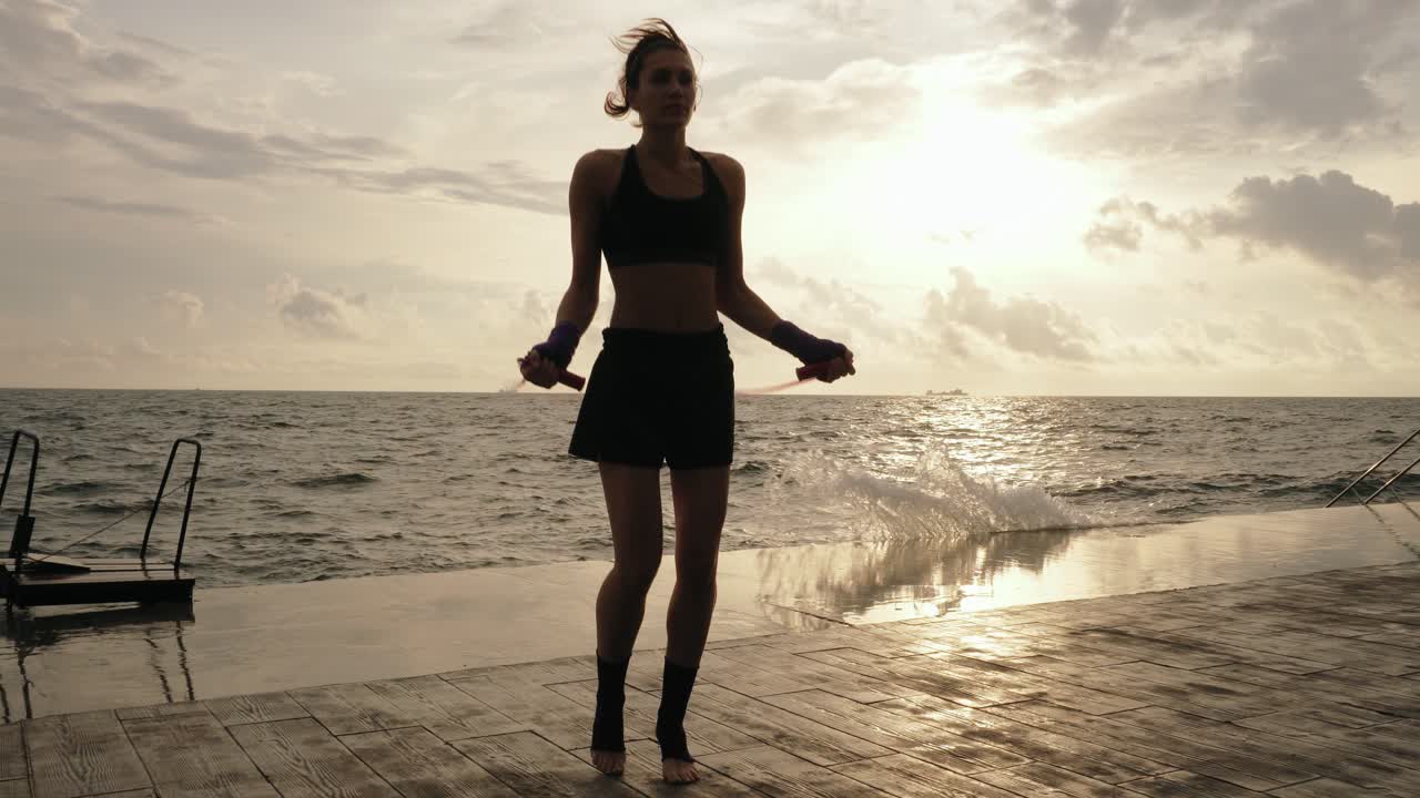Young woman working out on the jump rope against the sun by the beach in slowmotion. Lens flare. Girl jumping on a skipping rope by the sea. Shot in 4k