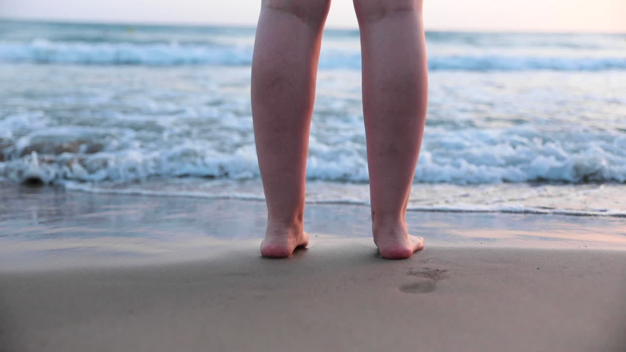 Piedi femminili nella sabbia accarezzati da un'onda durante un tramonto.