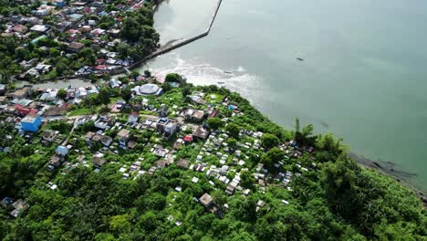 Vista-Aérea-De-Retroceso-Del-Cementerio-Tropical-Cubierto-De-Maleza-Frente-A-Las-Impresionantes-Aguas-De-La-Bahía-Del-Océano-Y-Al-Pueblo-Local