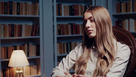 young woman psychologist listening to patient problems during therapy session sitting in comfortable armchair in library with bookshelves and lamp