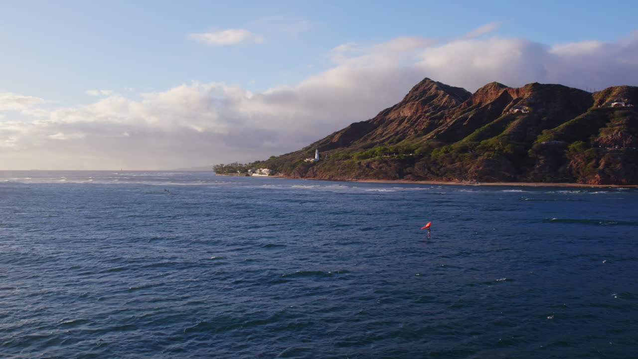 drone shot starting from Diamond head volcanic mountain formation on the island of Oahu in Hawaii across the Pacific to the sunset as kite boarders surf across the water