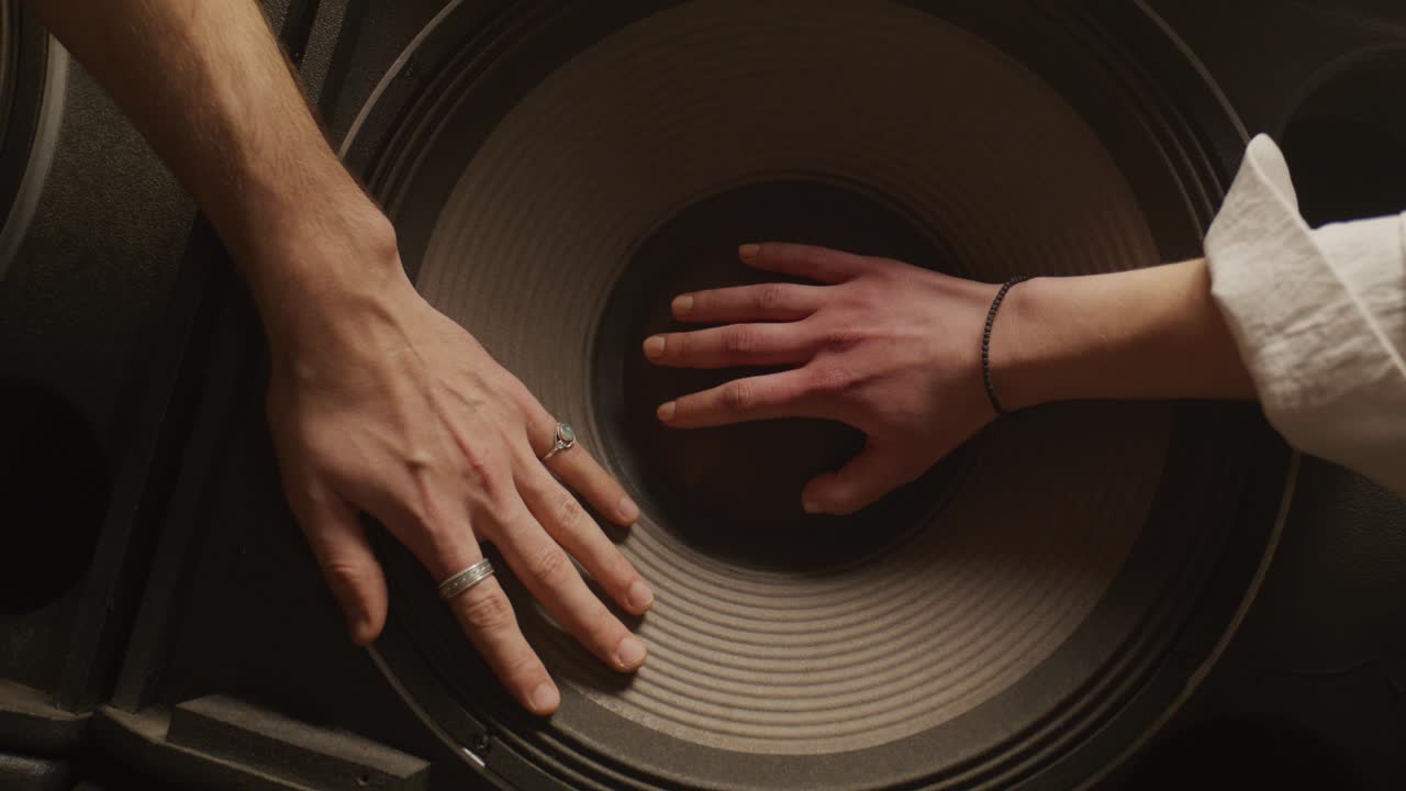 A top-down perspective of two people placing their hands on a large, vibrating subwoofer speaker, feeling the intensity of the bass