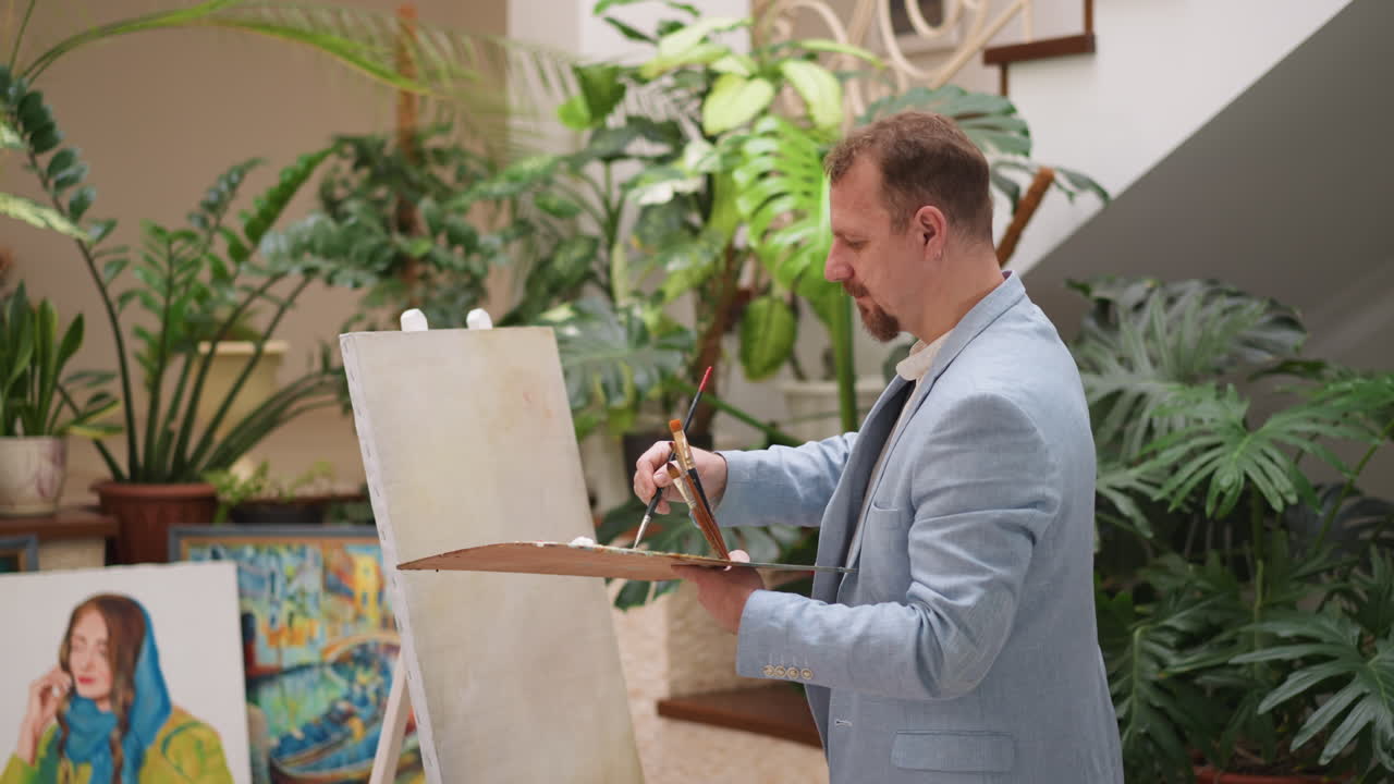 Artist with palette at empty canvas on easel. Bearded man waits for ideas holding slab and brushes set in sunny studio. Designer ready to draw masterpiece