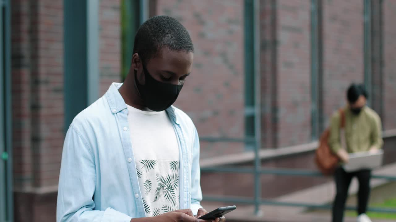 Camera zoom out on african american student wearing facial mask and using his smartphone near the college