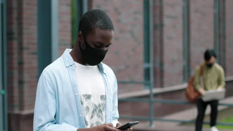 camera zoom out on african american student wearing facial mask and using his smartphone near the college