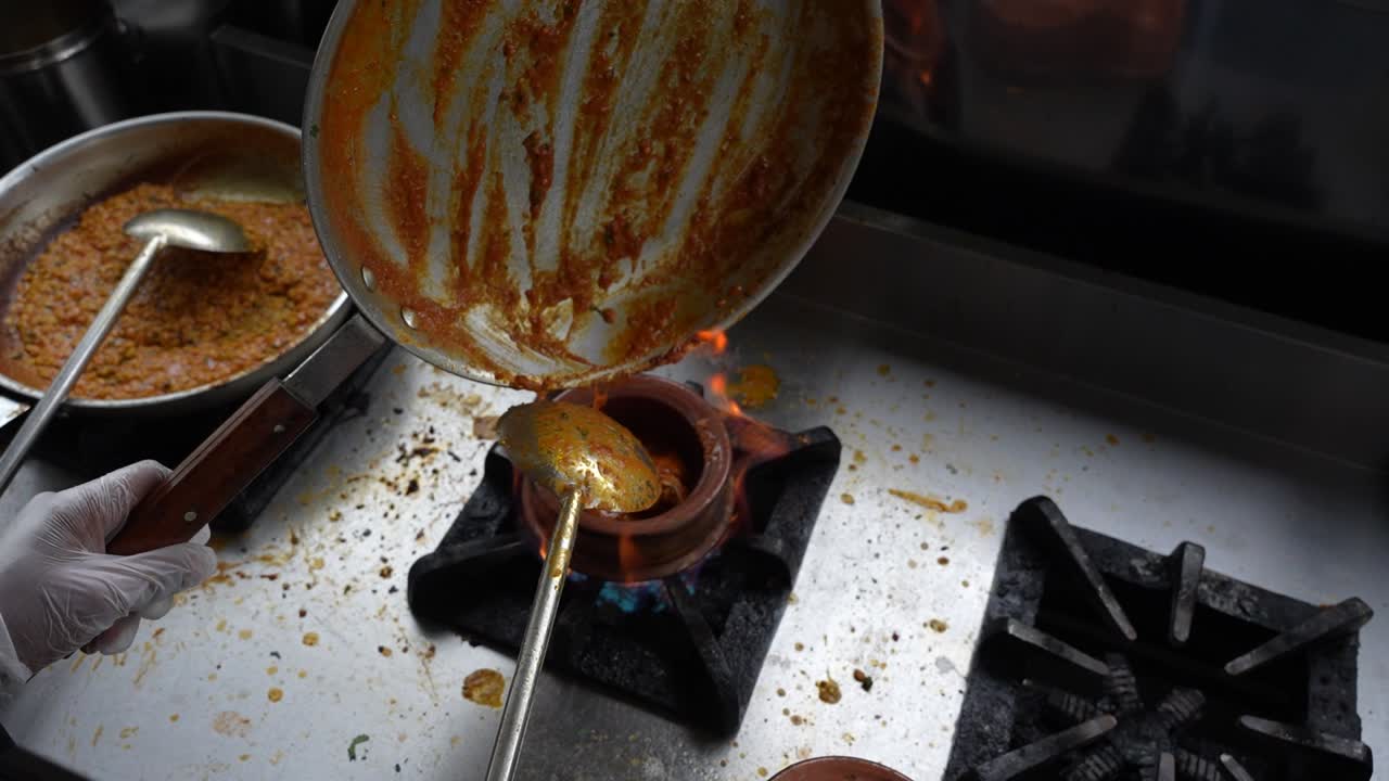 Chef transfers curry from a pan into a heated clay pot on a stove at a commercial kitchen