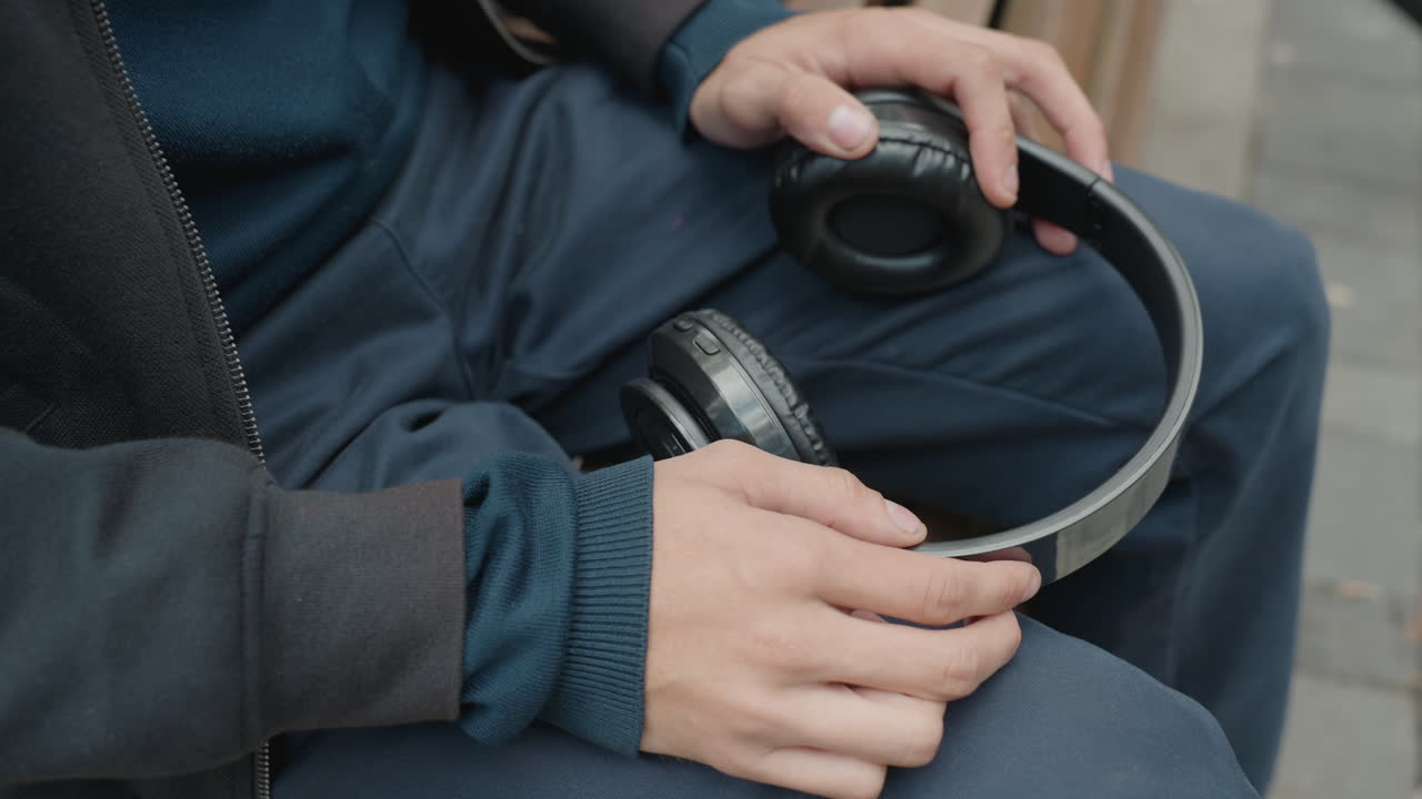 Mani che tengono delle cuffie sulle gambe, un pendolare si ferma su una panchina in città e regola il padiglione e l'imbottitura in pelle; i fotogrammi mostrano alternativamente pantaloni in denim e la manica di un maglione lavorato e un musicista di strada.