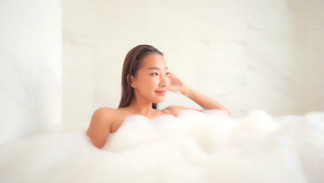 거품으로 가득 찬 욕조에서 스파에서 휴식을 취하고 창문을 바라보는 카메라를 바라보는 젊은 섹시한 아시아 여성, 슬로우 모션, 풀 프레임