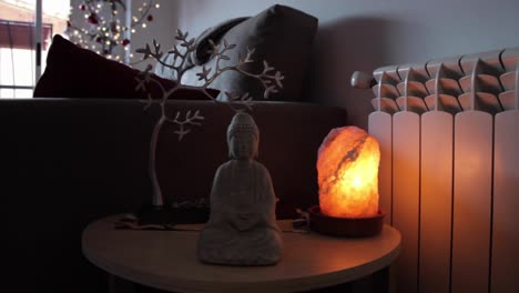 pequeña estatua de buda sentada en forma de flor de loto junto a una lámpara de piedra natural nale himalaya satl y un pequeño árbol de metal detrás en la sala de estar