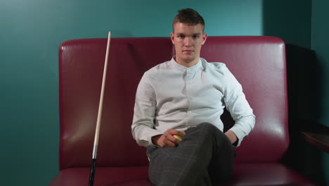 chap in white shirt and plaid pants sits on red sofa in billiard room, rolling orange billiard ball in hand. cue stick leans beside him. green wall background with stylish interior and dim lighting