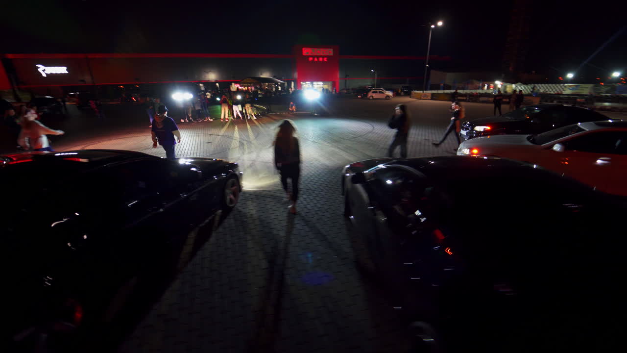Youngsters leave their cars driving seat and walking forward. Group of standing at backdrop. Square at night as backdrop.