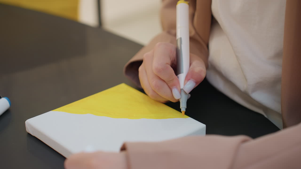 mano dibujando un lienzo vibrante con precisión, primer plano de una mano perfeccionando una obra de arte sobre una superficie amarilla, artista concentrado usando un rotulador para perfeccionar los detalles de un proyecto de arte colorido en un entorno de estudio sereno