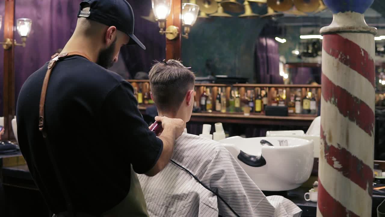 Back view of a young hairdresser cutting hair using electric trimmer on the neck of his customer in barber shot. Slow Motion shot