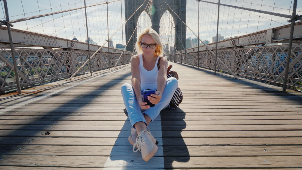 Una giovane donna seduta sul Ponte di Brooklyn a New York usa uno smartphone.