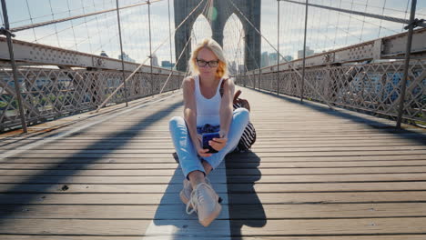 a young woman sits on the brooklyn bridge in new york uses a smartphone