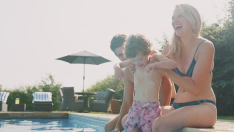 family with young son sitting on edge of pool having fun on summer vacation in outdoor swimming pool