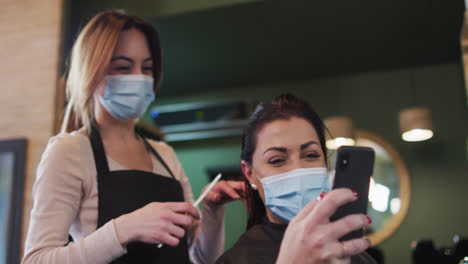 female hairdresser and female customer wearing face masks looking at smartphone at hair salon