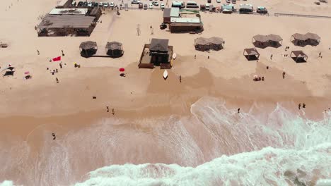 aerial-drone-showing-endless-wild-sea-waves-breaking-on-the-sandy-coast-line-while-people-hang-out-on-the-beach-and-a-life-guard-station-is-in-the-middle