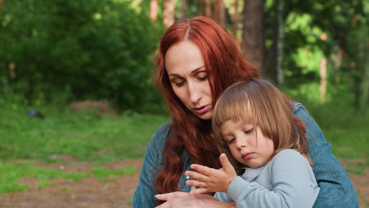 Mother carries little girl who looks unhappy while walking through green forest under soft daylight, showing tender care, emotional connection, and gentle parenting moment filled with love