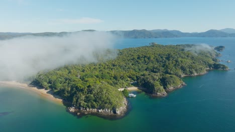 Rotating-aerial-view-of-remote-and-rugged-Ulva-Island,-part-of-Stewart-Island-Rakiura,-in-the-south-of-New-Zealand-Aotearoa,-with-moored-boat-in-secluded-white-sandy-beach-bay