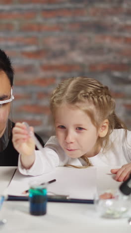 positive male asian teacher supports concentrated little girl dropping reagent into beaker with green liquid at fun science lesson in home lab