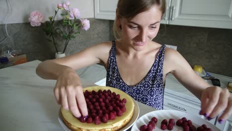 Young-smiling-woman-is-adoring-and-decorating-the-top-of-the-cake-with-raspberries-in-the-kitchen-at-home.-cheesecake-decorated