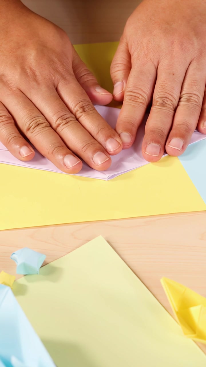 Mani adulte piegano carta origami pastello su una scrivania luminosa sotto un'illuminazione morbida e uniforme, vista dall'alto.