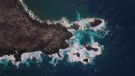 Aerial-top-down-of-whitewash-waves-from-deep-blue-ocean-water-crashing-on-eroded-cliff