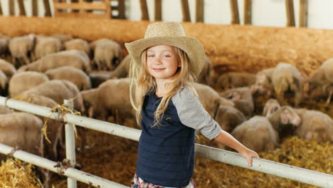 retrato de una niña caucásica con sombrero mirando y sonriendo a la cámara en un establo con rebaño de ovejas