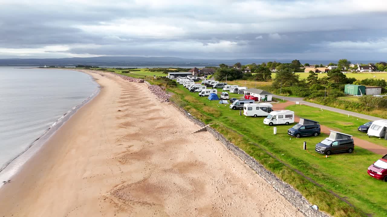 Drone footage glides along a coastal caravan park in Rosemarkie, Scotland, showing motorhomes, tents, and cars beside a sandy beach under cloudy daylight