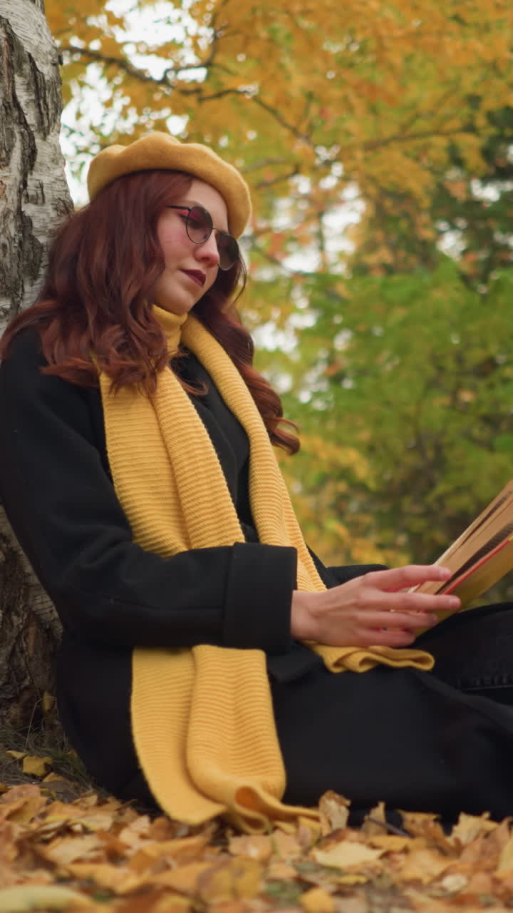 dama justa con boina amarilla se sienta en el suelo, cierra el libro, descansa la mano en él con una cálida sonrisa en un parque sereno, rodeado de follaje de otoño, disfrutando de un momento de paz en la naturaleza que irradia satisfacción