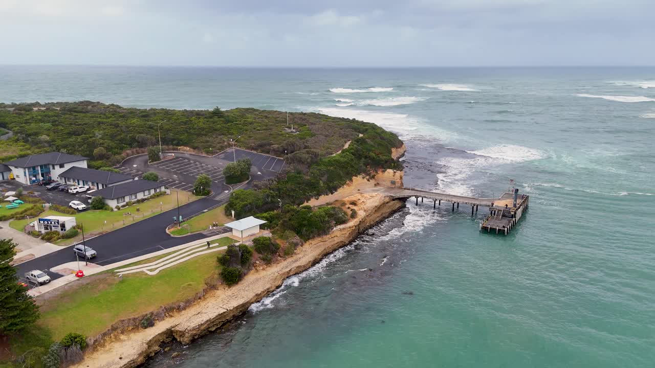 Drone captures sweeping views of Port Campbell's rugged coastline, pier, and lush greenery under overcast skies