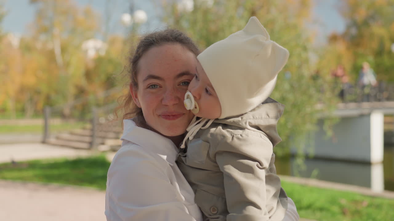 Madre che tiene in braccio il bambino nel parco. Luce solare autunnale che proietta un bagliore caldo, bacio spontaneo e coccole delicate, ciuccio in bocca al bambino, cappellino accogliente e cappotto imbottito. Sentiero lungo il lago e alberi verdi, gioioso legame familiare.