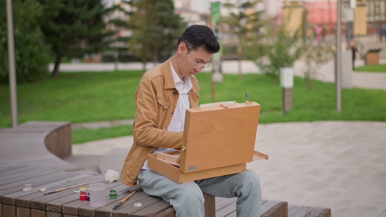 hombre asiático pintando en un banco del parque con un caballete portátil de madera y un set de acuarelas, botes y pinceles colocados en el banco, césped verde y senderos peatonales al fondo, práctica diurna, concentrado