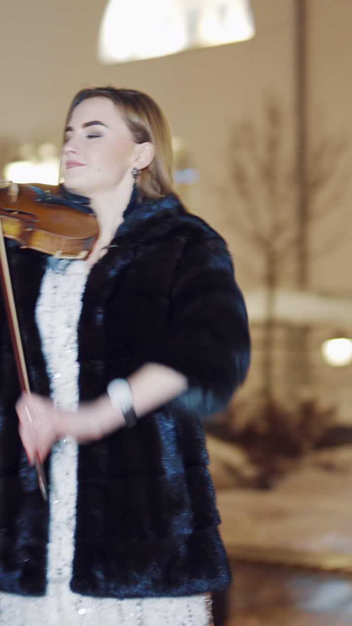 An attractive girl is standing in the square in a long white dress with sequins and playing the violin on the background of the monument and the buildings behind her in the evening. Blurred background. Vertical video