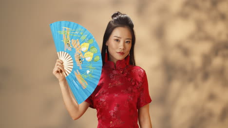 Retrato-De-Una-Joven-Asiática-Alegre-Vestida-Con-Ropa-Tradicional-Roja-Escondiendo-Su-Rostro-Con-Un-Abanico-Azul-Abierto