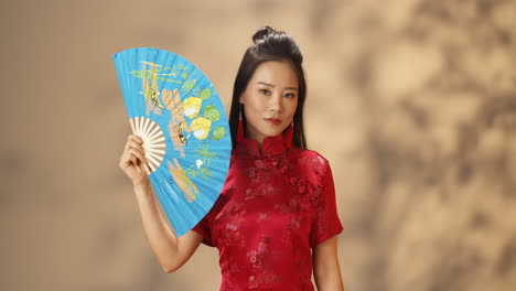 portrait d'une jeune femme asiatique joyeuse en vêtements traditionnels rouges cachant son visage avec un ventilateur bleu ouvert