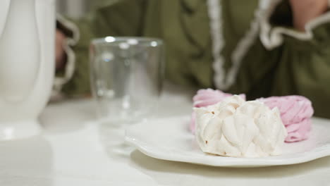 close up of creamy dessert on plate with woman placing glass cup gently on table while sitting down in bright cozy kitchen, capturing soft moment of tea time preparation