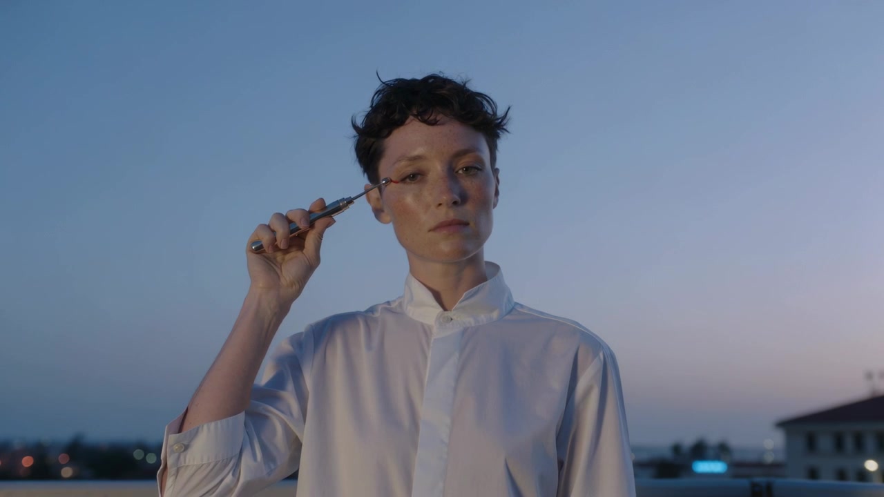 Una donna in camicia bianca, tenendo lo scovolino, applica il mascara lungo le ciglia destre su un tetto al tramonto per il trucco.