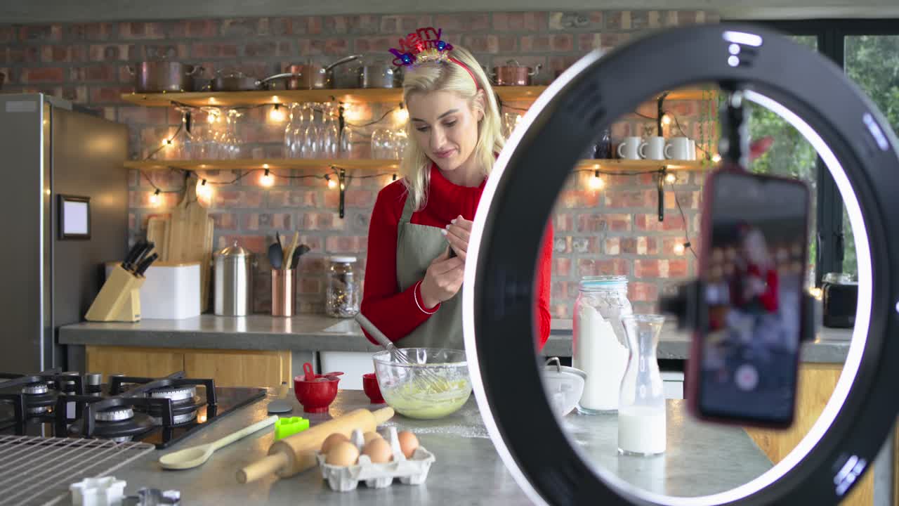 Woman whisking batter at kitchen island facing smartphone camera then adding vanilla extract