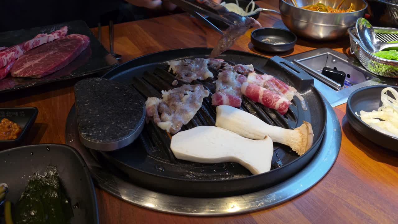 Assorted meats and king oyster mushrooms grilling on a tabletop Korean BBQ grill with side dishes around