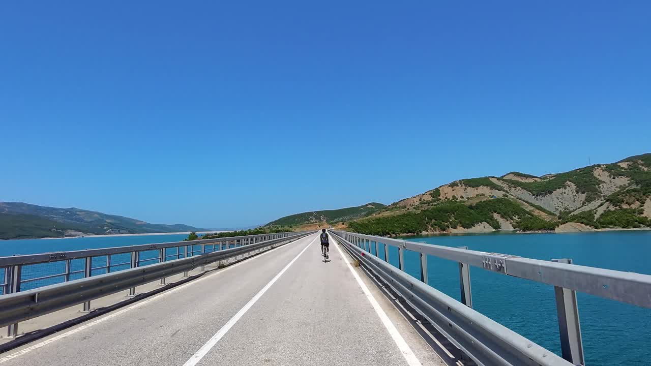 Happy Cyclist Riding on Long Bridge Over Blue Lake Fjords Road in Scenic Albanian Hills